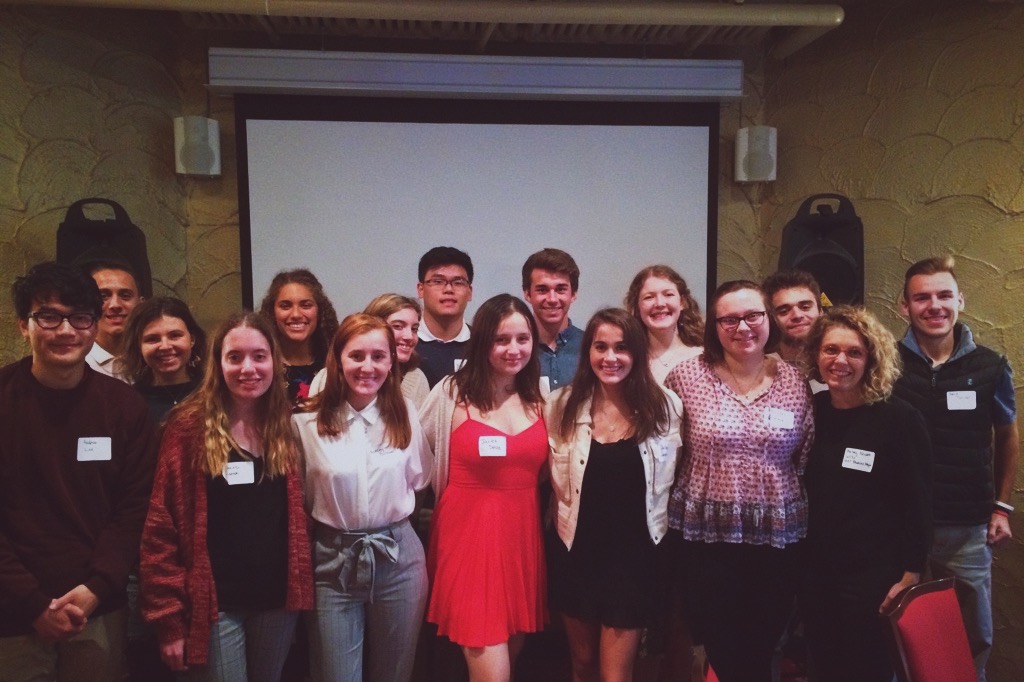 16 people posing for a photo. projector screen and speakers in the background.